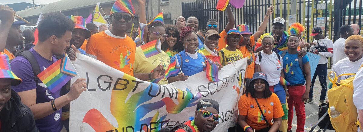 A group of smiling and happy people in colourful clothes holding a banner saying 'LGBT Unity Scotland, End Detention' and with a rainbow flag waving.